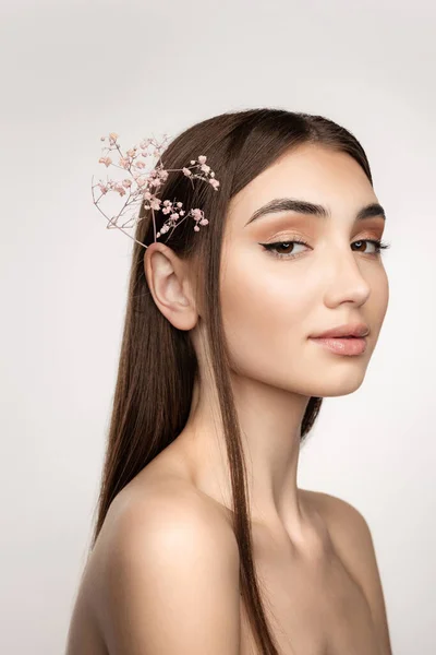 Gyönyörű fiatal lány egyenes barna haj és gyengéd smink úgy néz ki, pózol a virág a haj elszigetelt szürke háttér — Stock Fotó