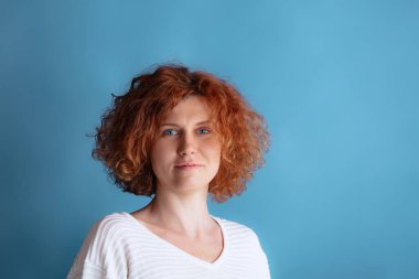 Kıvırcık saçlı, parlak mavi gözlü güzel bir kadının portresi. Mavi stüdyo arka planında izole edilmiş poz veriyor.