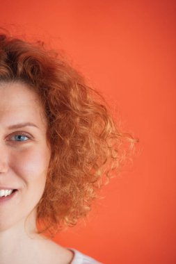 Mavi gözlü, kıvırcık saçlı, gülümseyen, kırmızı stüdyo arka planında izole bir şekilde poz veren güzel kızıl saçlı kadın.