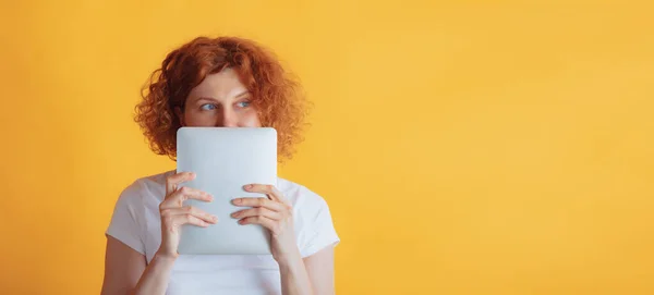 Portrait of young red-haired woman covering face with tablet, thinking, poisng isolated over yellow studio background — Fotografia de Stock