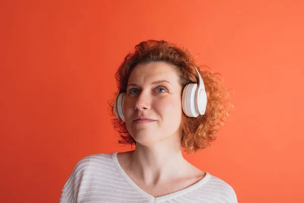 Portrait of red-haired woman having online lesson, listening to infromation in headphones isolated over red studio background — 스톡 사진