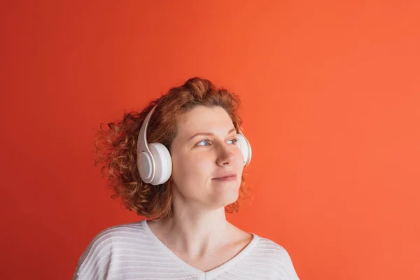 Portrait of calmly looking red-haired woman listening to music in white headphone isolated over red studio background — 스톡 사진