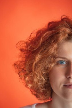 Mavi gözlü, kıvırcık saçlı, kızıl saçlı güzel bir kadının yarım yüzlü portresi, sakince kameraya bakıyor, kırmızı stüdyo arka planında izole edilmiş poz veriyor.