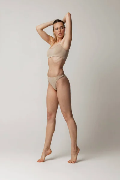 Полноразмерный портрет нежной молодой женщины с идеальной формой тела, позирующей в бежевом хлопковом нижнем белье, изолированном на сером фоне студии — стоковое фото