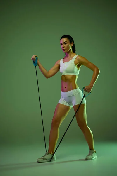 Porträt eines jungen muskulösen Mädchens beim Training, das Handübungen mit speziellen Sportgeräten macht, isoliert über grünem Studiohintergrund in Neonlicht — Stockfoto