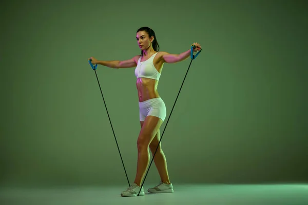 Full-length portrait of young sportive girl training, doing exercises isolated over green studio background in neon light — Foto de Stock