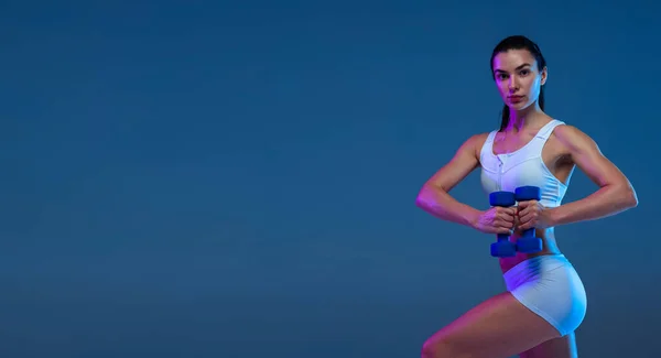 Half-length portrait of young sportive girl training, doing exercises with dumbbell isolated over blue studio background in neon light — Foto de Stock