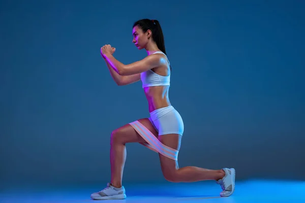 Portrait of young sportive woman training, doing squats with fitness elastic band isolated over blue studio background in neon light — Stok fotoğraf