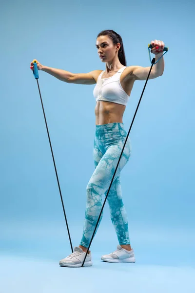 Full-length πορτρέτο των νεαρών σπορ κορίτσι κατάρτισης, κάνει ασκήσεις με αθλητικό εξοπλισμό απομονώνονται σε μπλε φόντο στούντιο. — Φωτογραφία Αρχείου