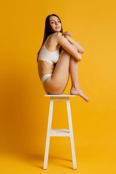 Ніжна молода дівчина сидить на стільці, позує в нижній білизні ізольовано на жовтому студійному фоні — стокове фото