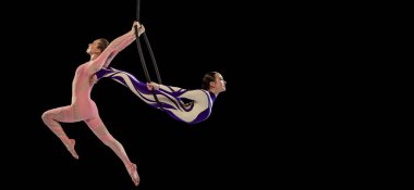 Portrait of two young girls, professional air gymnasts performing on hoop isolated over black studio background. Flyer
