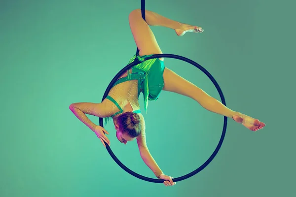 Portrait of young girl, professional air gymnast training on hoop isolated over green background in neon light — стоковое фото