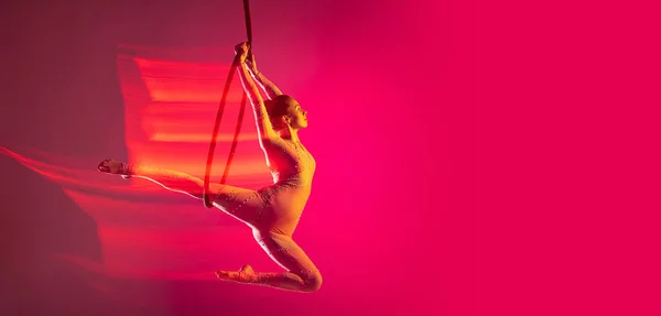 Dynamic portrait of young artistic girl, professional air gymnast training, performing isolated over pink background with mixed lights effect. Flyer — Stok fotoğraf