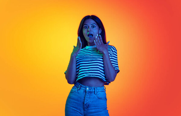 Portrait of young girl with shocked facial expression isolated over gradient orange yellow studio background in neon light