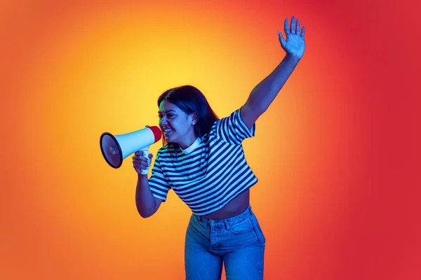 Portrait of young girl shouting in megaphone, posing isolated over gradient red yellow background in neon light — Fotografia de Stock