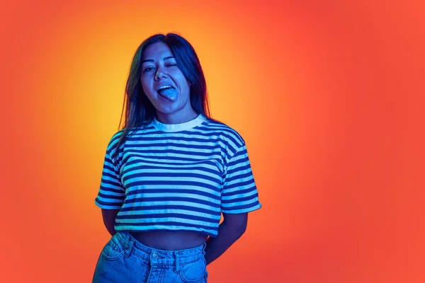 Egy fiatal, érzelmes lány portréja, akinek a nyelve narancssárga háttér felett áll ki a neon fényben. Vicces arc. — Stock Fotó