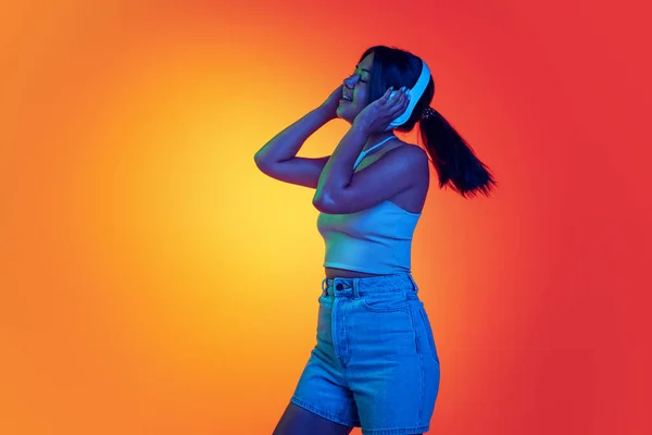Fiatal vidám nő hallgat zenét fülhallgató elszigetelt felett gradiens sárga narancs háttér neon fény — Stock Fotó