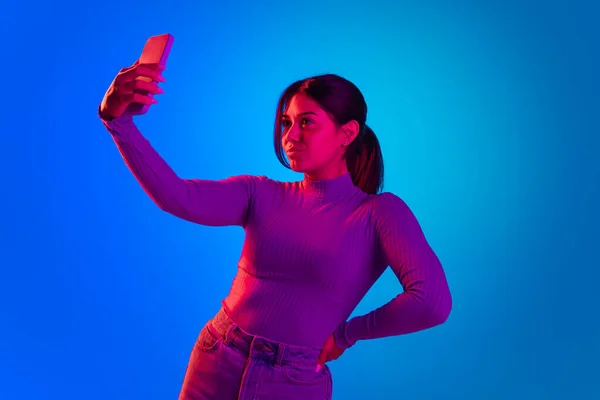Young beautiful girl taking selfie with phone front camera isolated over blue background in pink neon light — 스톡 사진