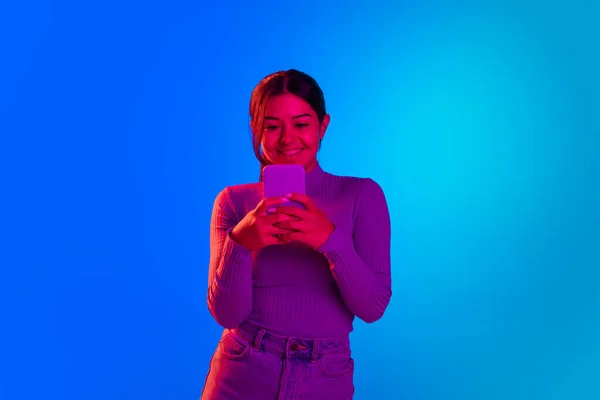 Portrait of young cheerful girl reading text messages on phone, smiling isolated over blue background in neon light — 스톡 사진