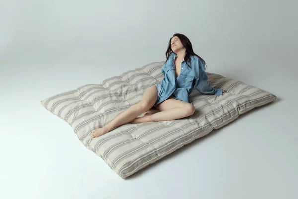 Una hermosa joven sentada en el colchón, posando en ropa interior y camisa aislada sobre fondo gris del estudio. Belleza natural —  Fotos de Stock