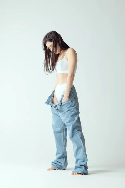 Full-length portrait of young slim woman wearing big jeans, posing isolated over grey studio background. Weight-loss concept — Stockfoto