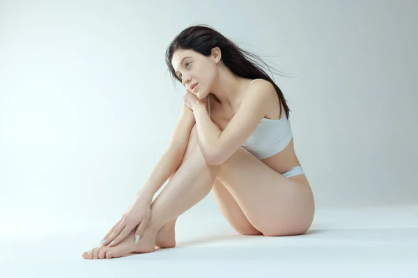 Mujer joven tierna sentada en el suelo, posando en ropa interior aislada sobre fondo gris del estudio. Atención médica femenina —  Fotos de Stock