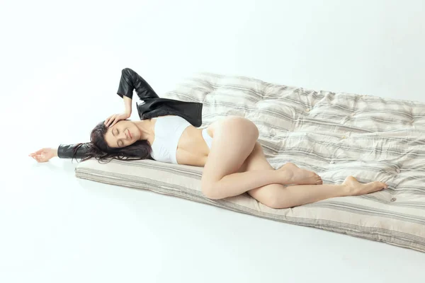 Одна красивая стройная девушка лежит на матрасе в белом нижнем белье и черной куртке, изолированной на сером фоне студии. Комфортный отдых — стоковое фото