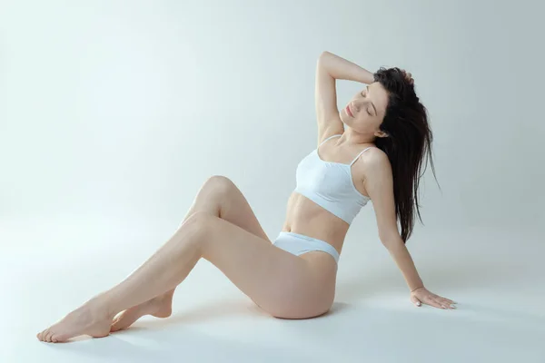 Portrait of young beautiful woman posing in white cotton underwear, sitting on floor isolated over grey studio background — Stock Fotó