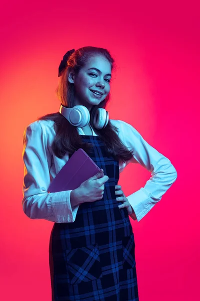 Porträt des netten jungen Mädchens, Studentin in Hemd und kariertem Kleid posiert mit Tablet isoliert über rosa Studiohintergrund im Neonlicht — Stockfoto