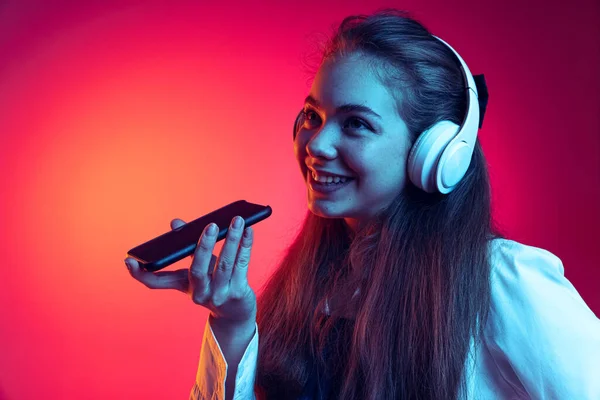 若い陽気な女の子の肖像,ネオンライトでピンクのスタジオの背景に孤立電話で音声メッセージを録音ヘッドフォンの学生 — ストック写真