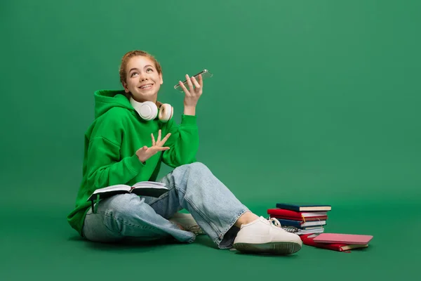 若い女の子の肖像画、カジュアルな布で学生、床に座って、陽気に電話で緑のスタジオの背景に隔離された話 — ストック写真