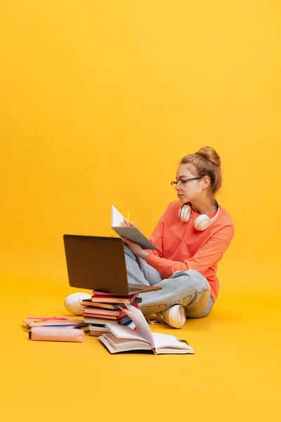 Retrato de menina, estudante de camisola laranja, óculos sentados no chão, fazendo anotações, estudando isolado sobre fundo amarelo — Fotografia de Stock