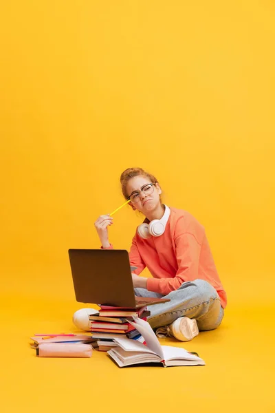 Retrato de menina, estudante de camisola e óculos sentados no chão com expressão pensativa, estudando isolado sobre fundo amarelo — Fotografia de Stock