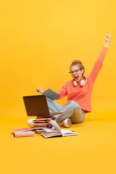 Jovem, estudante de óculos e suéter sentado no chão com laptop, estudando isolado sobre fundo amarelo. Tenho a maior marca. — Fotografia de Stock