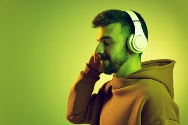 Bıyıklı ve piercingli genç bir adamın portresi yeşil stüdyo arka planında izole edilmiş kulaklıkla müzik dinliyor.