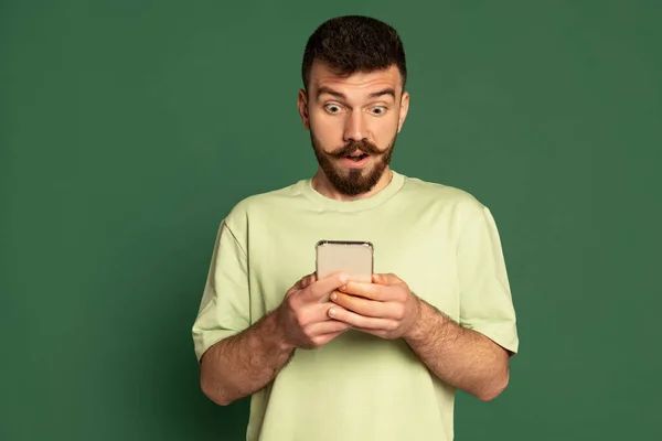 Portrait de jeune homme émotif regardant le téléphone avec une expression choquée isolé sur fond de studio vert. Message expressif — Photo