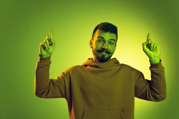 Retrato de hombre joven y elegante en sudadera casual posando con sonrisa aislada sobre fondo de estudio verde en luz de neón —  Fotos de Stock