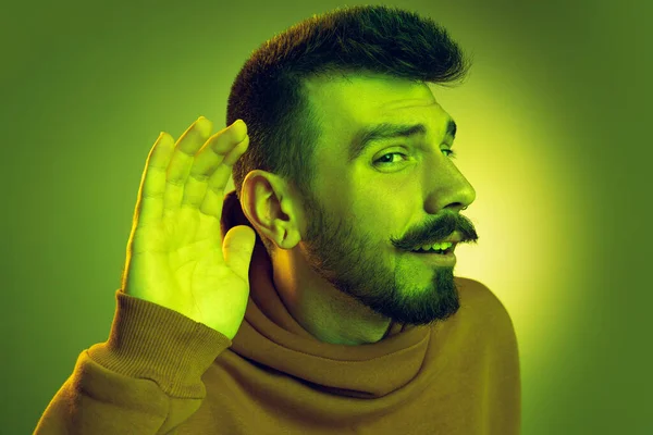 Retrato de jovem com a mão ao ouvido posando isolado sobre o fundo do estúdio verde em luz de néon. Audiência — Fotografia de Stock