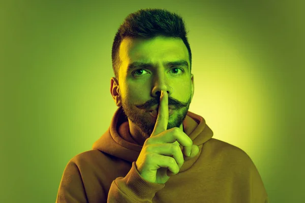 Πορτρέτο του νεαρού άνδρα βάζοντας δάχτυλο στα χείλη, θέτοντας απομονωμένη πάνω από το πράσινο φόντο στούντιο — Φωτογραφία Αρχείου