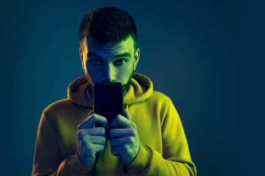 Cep telefonu tutan genç adamın portresi, kameraya bakıyor, neon ışıkta mavi arkaplanda izole bir şekilde poz veriyor.