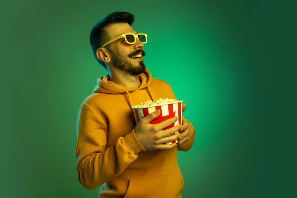 Portré egy szemüveges fiatalemberről, popcorn dobozzal, zöld háttérrel elszigetelve, neon fényben. Nevetés — Stock Fotó