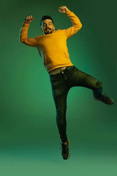 Ολόσωμο πορτραίτο του νεαρού άνδρα με casual cloth jumping, να ποζάρει απομονωμένος πάνω από πράσινο φόντο στούντιο σε νέον φως. Ενθουσιασμός — Φωτογραφία Αρχείου