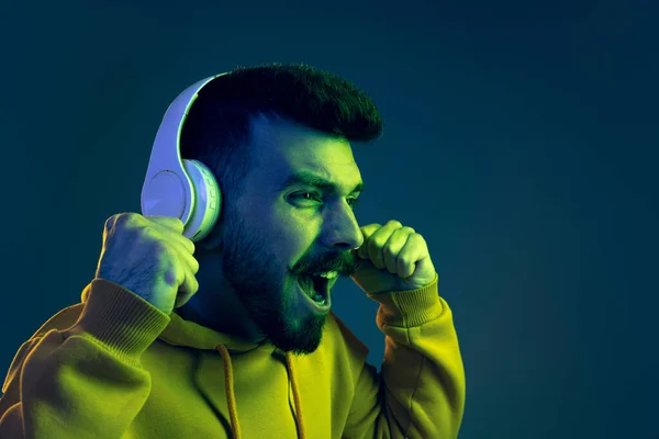 Portrait of young emotive man listening to dynamic music in headphones isolated over blue background in neon light — Stock Photo, Image