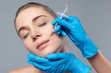 Gri stüdyo arka planında izole edilmiş dudaklara kozmetoloji enjeksiyonu yapan genç kızın portresi. Dudak büyütme