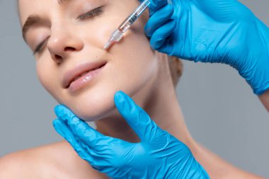 Gri stüdyo arka planında izole edilmiş dudaklara kozmetoloji enjeksiyonu yapan genç kızın portresi. Dudak büyütme