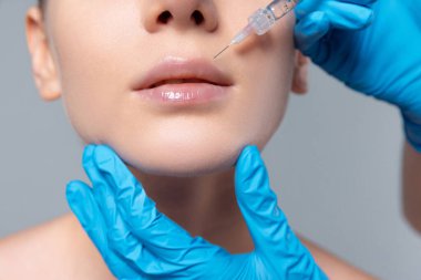 Gri stüdyo arka planında izole edilmiş dudaklara kozmetoloji enjeksiyonu yapan genç bir kız görüntüsü. Dudak büyütme.