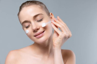 Cildine özen gösteren güzel bir kadının portresi. Gri stüdyo arka planına izole edilmiş nemlendirici krem sürüyor.