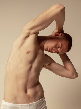 Gri stüdyo arka planında izole edilmiş beyaz iç çamaşırlarıyla poz veren genç kızıl saçlı yakışıklı adam portresi. Kas gevşetici vücut. Erkeklik. Erkeklerin sağlık, güzellik, vücut ve cilt bakımı, uygunluk kavramı.