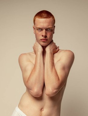 Gri stüdyo arka planında çıplak poz veren kızıl saçlı genç adamın portresi. Çilli vücut. Kameraya ciddi ciddi bak. Erkek güzelliği kavramı. Refah, spor, kabullenme, moda
