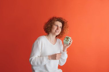 Kutlama. Kırmızı stüdyo arka planında izole edilmiş küçük hediye kutusunu tutan mutlu görünümlü kızıl saçlı kadının portresi. Doğum günü hediyesi. Yüz ifadesi kavramı, duygular, duygular, reklamlar